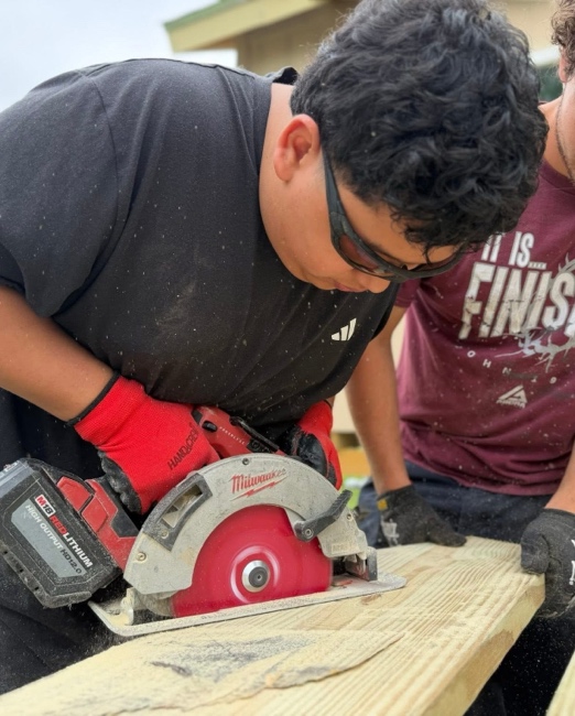 A young AGORA Construction student in safety glasses cutting lumber with a circular saw beside a mentor